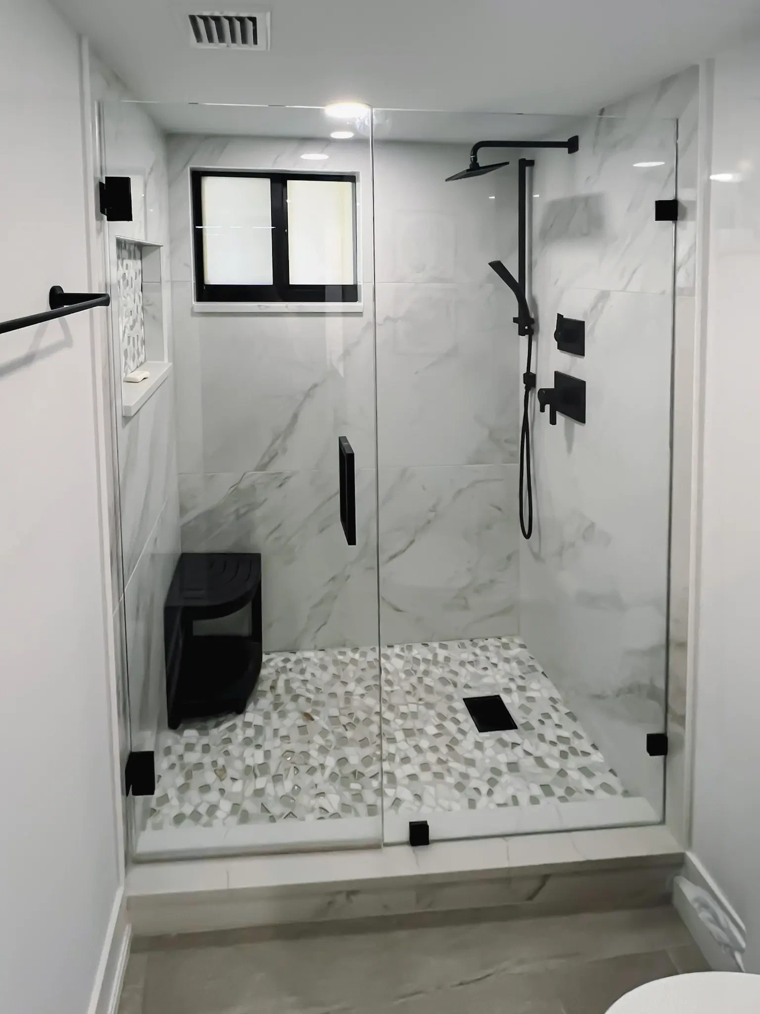 Modern black-framed glass shower with marble walls, pebble floor, and sleek fixtures for luxury bathroom design.