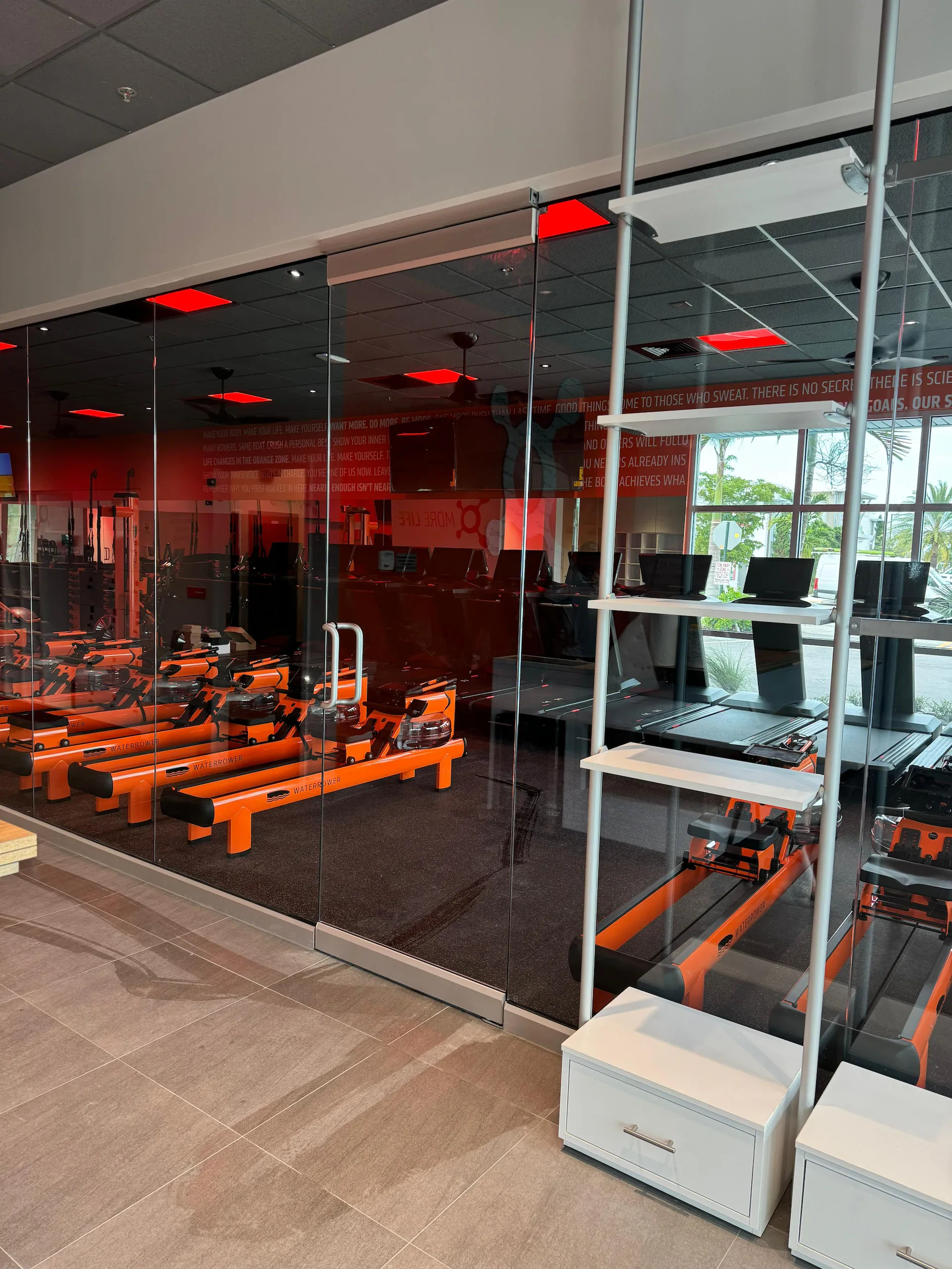 Reflective glass walls in a modern gym with orange workout equipment and computer stations, showcasing custom mirror installations.
