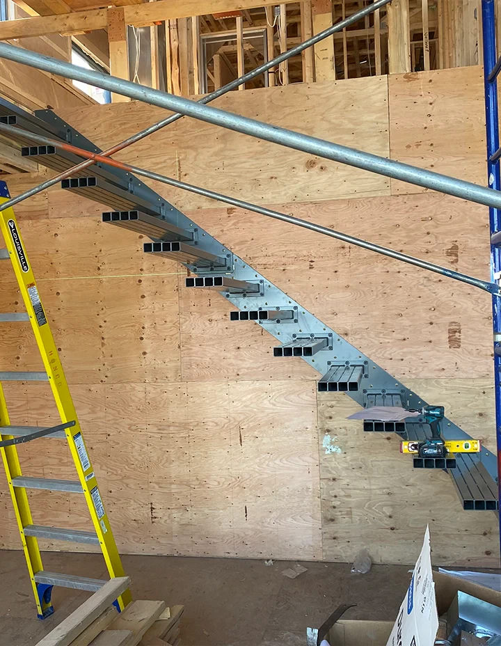 Image of a steel-framed glass staircase under construction at a building site, surrounded by scaffolding and wooden wall panels.