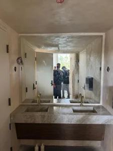 Mirror reflection showing double marble sinks, gold fixtures, and construction workers in a stylish bathroom setting.