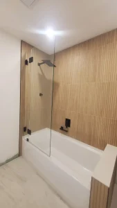 Modern glass shower enclosure with black hardware and wood-textured tiles.