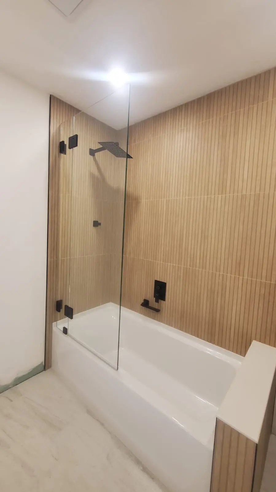 Modern glass shower enclosure with black hardware and wood-textured tiles.