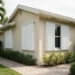 White accordion hurricane shutters closed over the windows of a South Florida house with one shutter left half open
